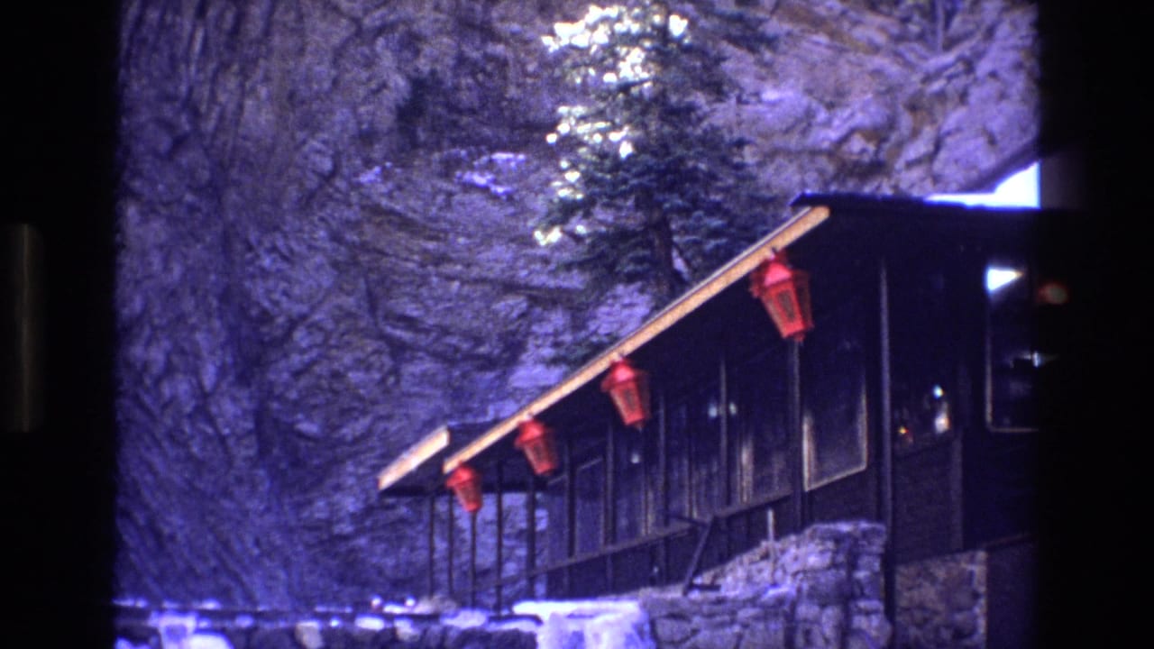 1973 Colorado 8mm frame with the cabin seen from the side, red lanterns under the roofline, snow at the base, and a rugged rock wall in the background.