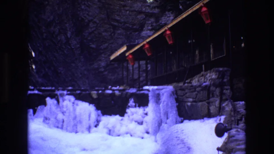 1973 Colorado 8mm frame showing the cabin partly in shadow with red lanterns under the eaves, deep snow, icicles, and a rocky winter setting.