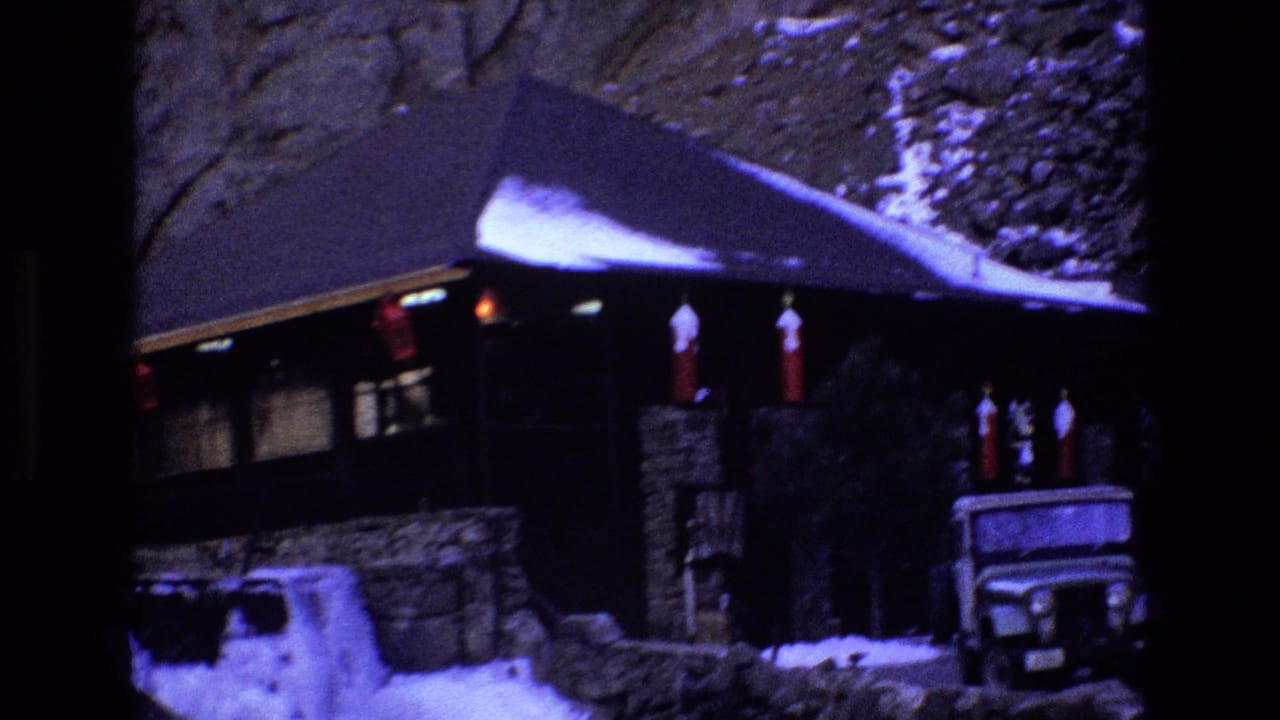 1973 Colorado 8mm frame showing a snow-dusted stone-and-wood mountain cabin with red lanterns under the eaves and a Jeep-style vehicle parked near the entrance.