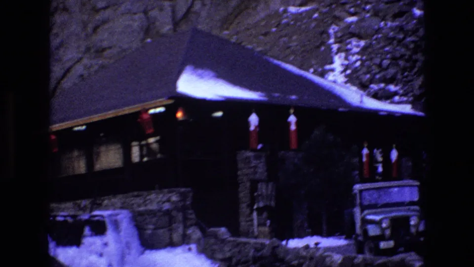 1973 Colorado 8mm frame showing a snow-dusted stone-and-wood mountain cabin with red lanterns under the eaves and a Jeep-style vehicle parked near the entrance.