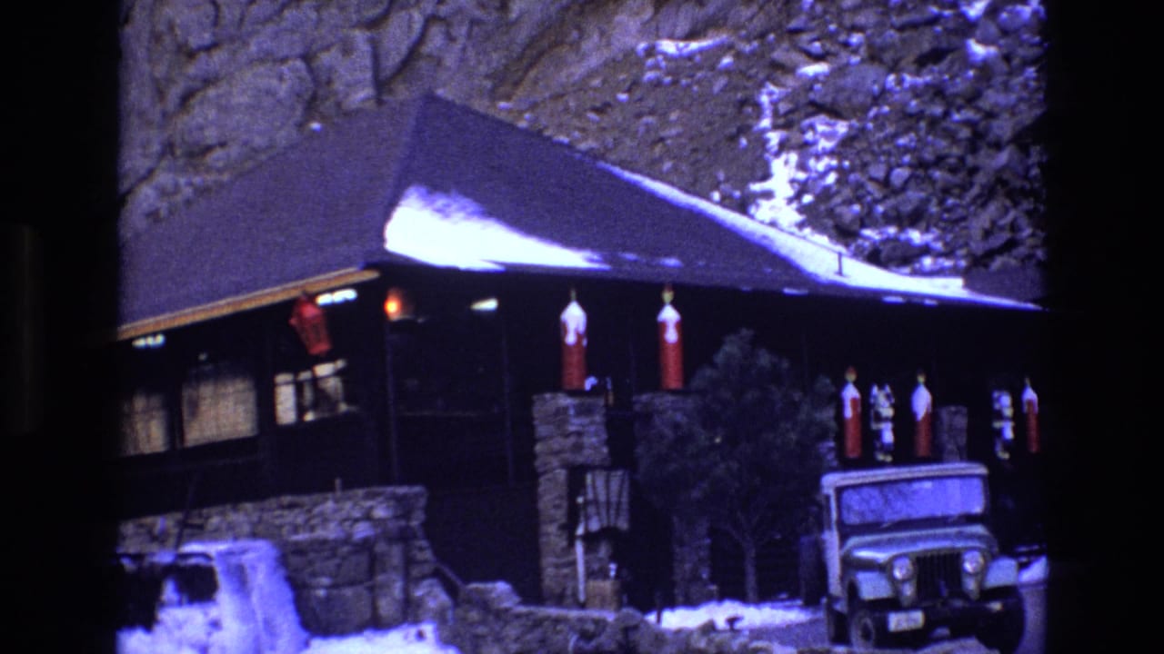 1973 Colorado home-movie frame of a rustic cabin or lodge exterior, with snow along the roofline, red lanterns, stonework, and a Jeep-style vehicle in front.