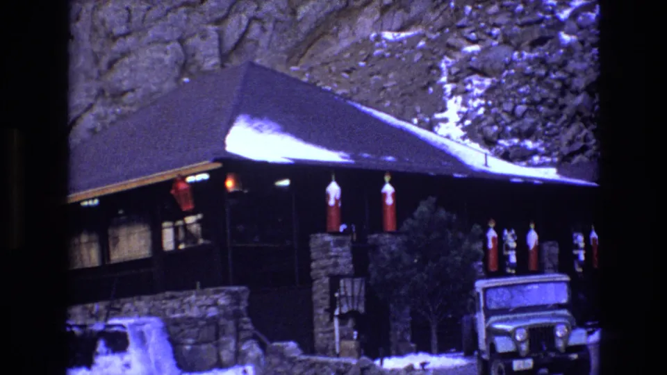 1973 Colorado 8mm frame with the cabin filling the view, showing a snowy sloped roof, hanging red lanterns, stone supports, and the front of a Jeep-style vehicle.