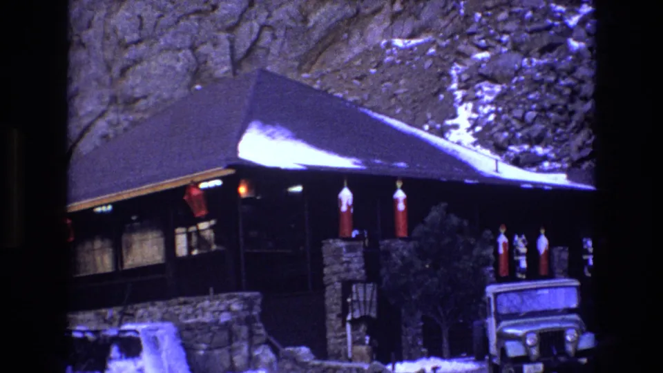 1973 Colorado home-movie frame showing the same snowy mountain cabin exterior with red lanterns, stonework, shrubs near the entrance, and a Jeep-style vehicle parked outside.