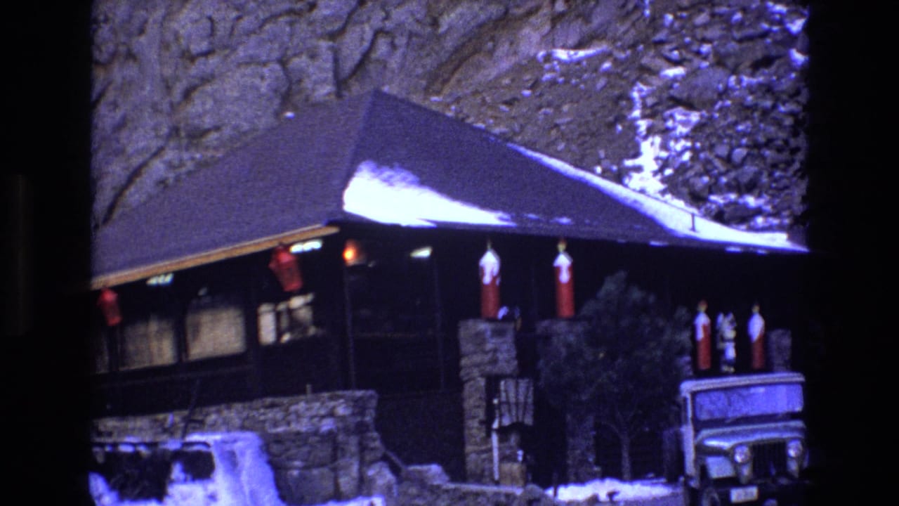 1973 Colorado 8mm frame showing a dark wood-and-stone mountainside cabin with snow on the roof, red exterior decorations, and a white Jeep-style vehicle partly visible in front.