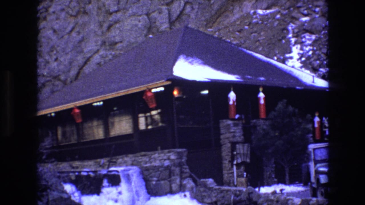 Cabin exterior with snow, red decorations, stonework, and a partially visible parked vehicle.