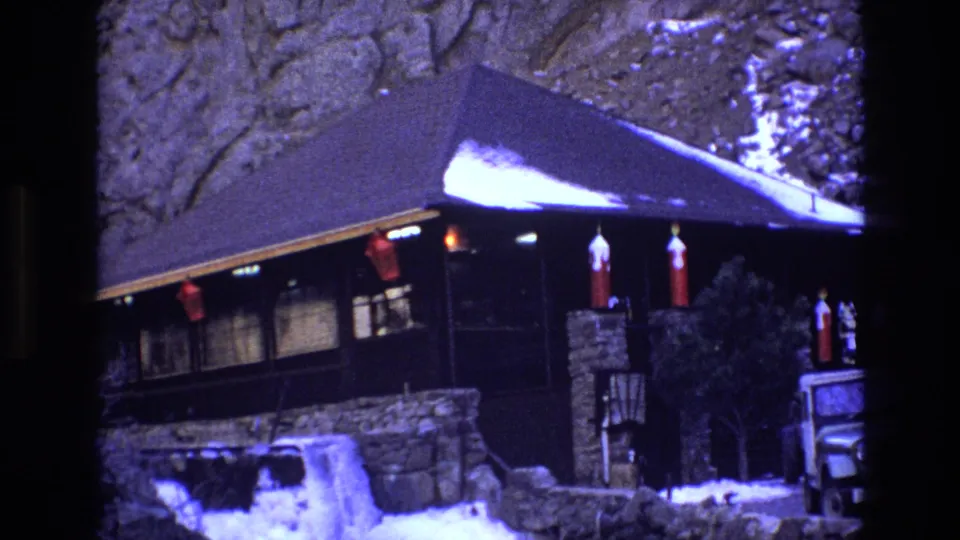1973 Colorado 8mm frame showing the mountainside cabin from a similar angle, with a snow-dusted roof, stone retaining wall, red decorations, and a white Jeep-style vehicle at the lower right.