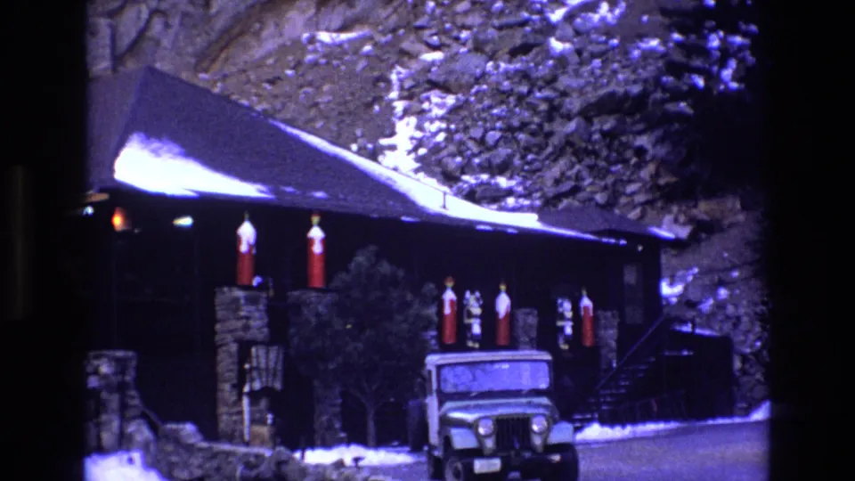 1973 Colorado home-movie frame with a wider view of the dark mountainside cabin, snow along the roof and ground, red exterior decorations, small figures, and a white Jeep-style vehicle in front.