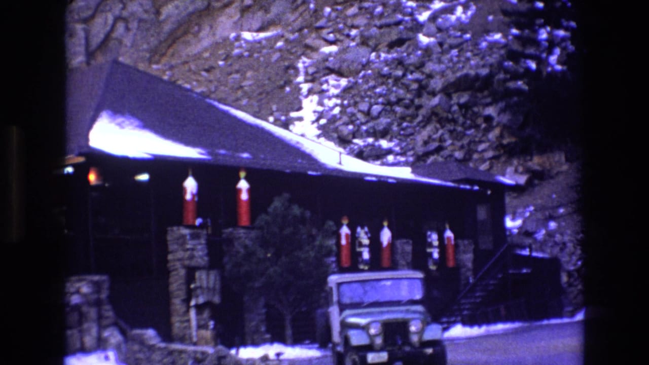 1973 Colorado 8mm frame showing a rustic dark-wood cabin below a rocky snowy slope, with red holiday decorations and a green Jeep-like vehicle near the entrance.