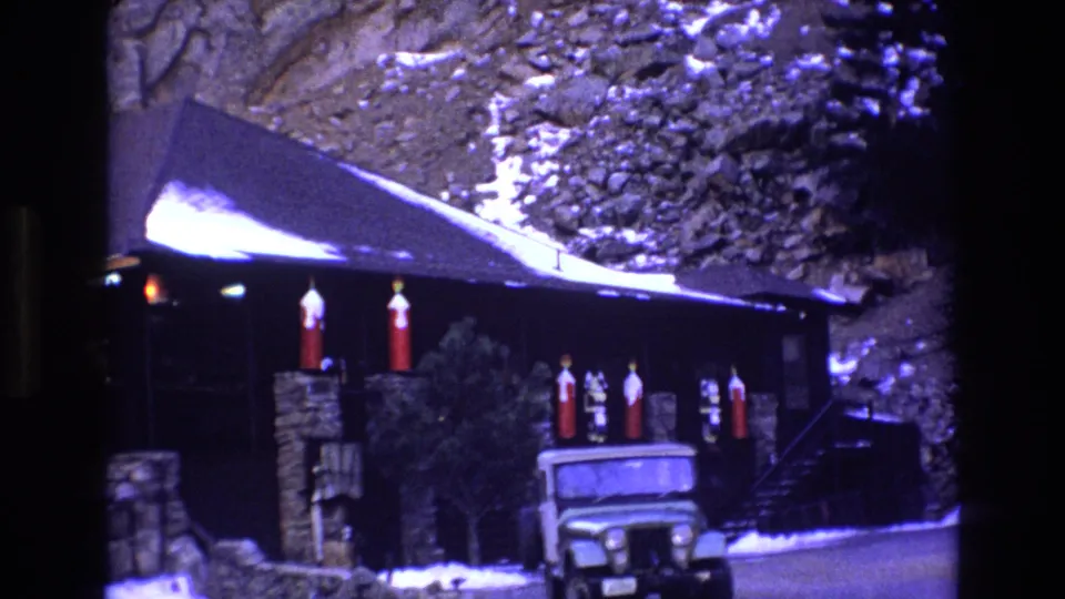 1973 Colorado 8mm frame showing a rustic dark-wood cabin below a rocky snowy slope, with red holiday decorations and a green Jeep-like vehicle near the entrance.