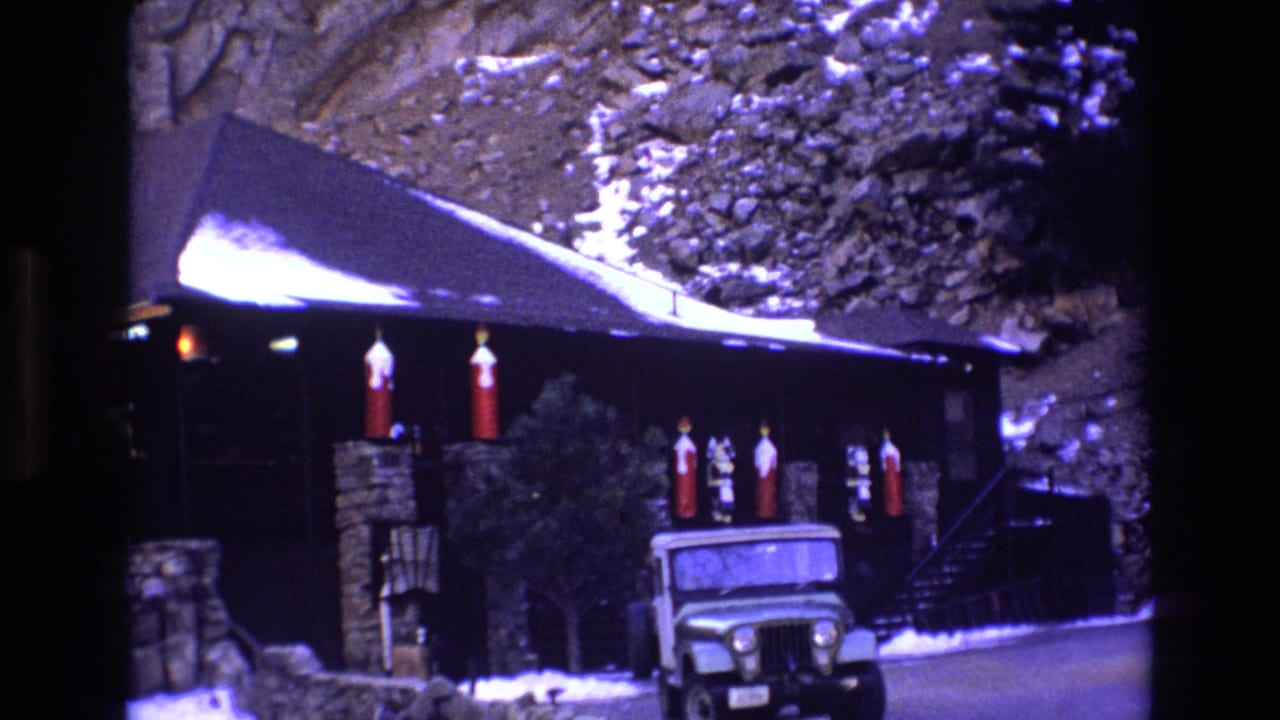 Rustic snowy cabin exterior with stone posts, red decorations, and a parked green vehicle.
