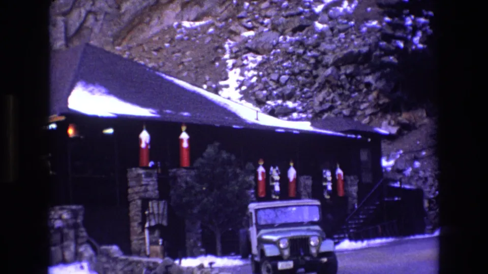 1973 Colorado home-movie frame of a snow-lined cabin front with stone supports, red holiday decorations, exterior stairs, and a green Jeep-like vehicle below the porch area.