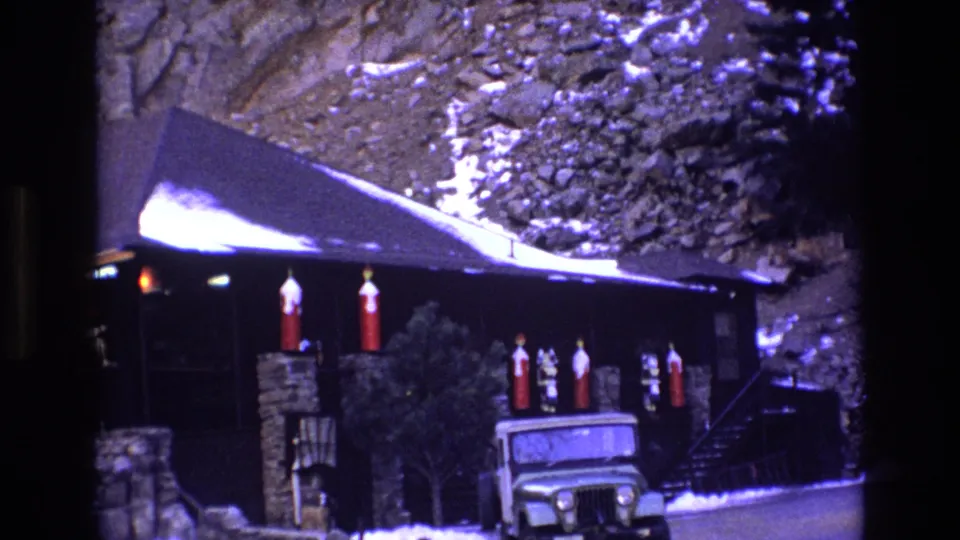 1973 Colorado 8mm frame holding on the alpine cabin exterior, with snow along the roofline, red holiday ornaments, a green Jeep-like vehicle, and a steep rocky slope behind it.
