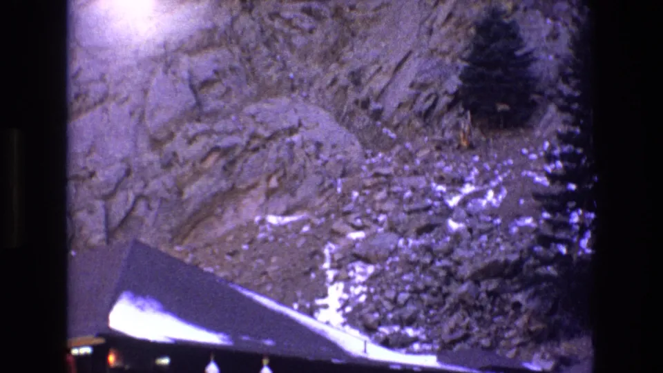 1973 Colorado home-movie frame shifting upward from the cabin to the surrounding rocky mountainside, with scattered snow patches and evergreen trees visible above the roofline.