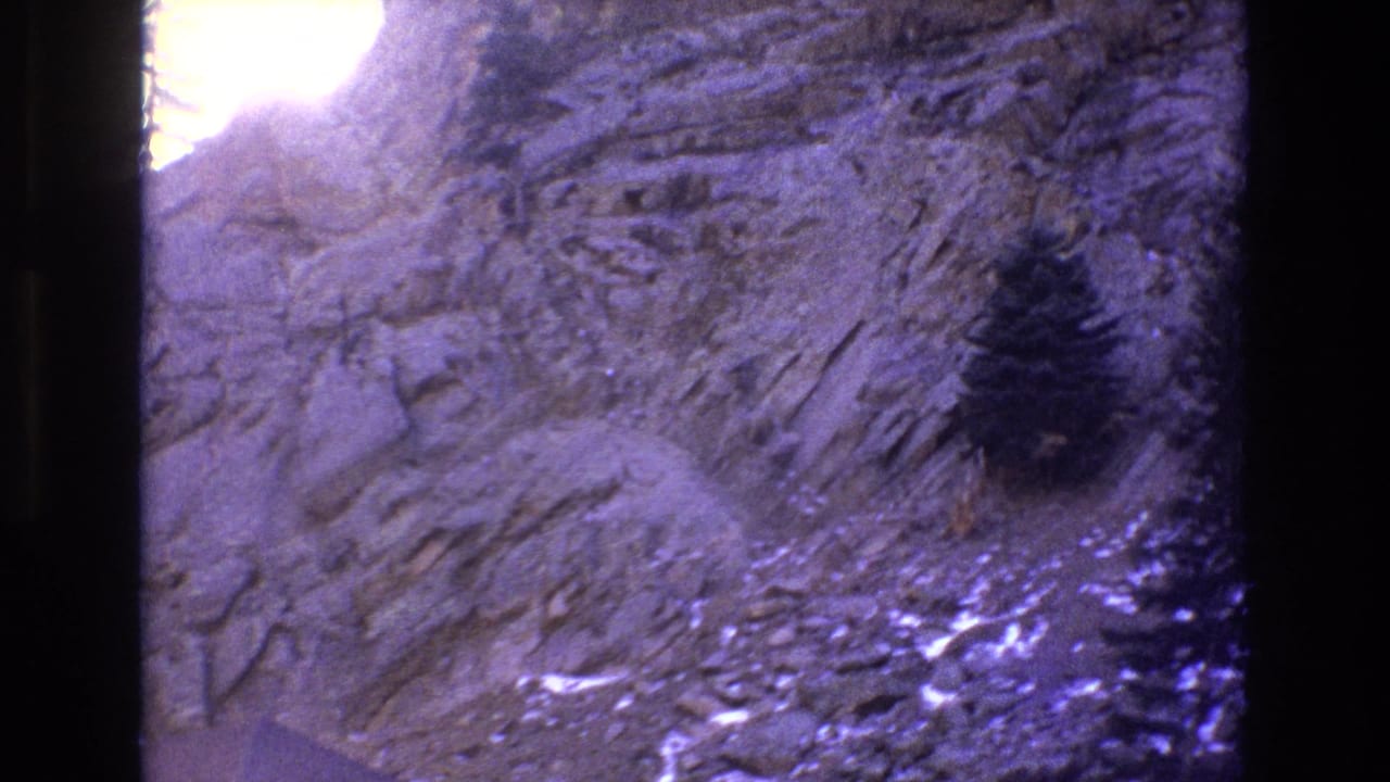1973 Colorado 8mm frame showing a stratified rocky slope, a small dark evergreen, and bright sunlit overexposure along the upper-left edge.