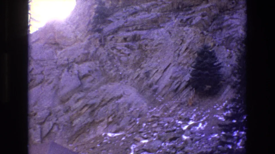 1973 Colorado 8mm frame showing a stratified rocky slope, a small dark evergreen, and bright sunlit overexposure along the upper-left edge.