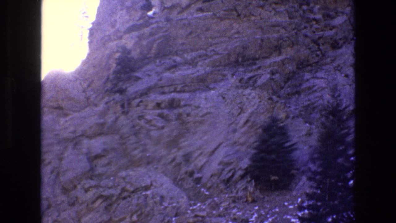 1973 Colorado 8mm frame with a steep rocky cliff face, dark evergreen growth, and blown-out sunlight near the upper-left ridge line.