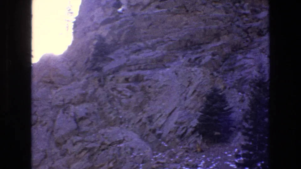 1973 Colorado 8mm frame showing a darker exposure of rugged rock strata, a small evergreen, and a bright overexposed opening along the left side.