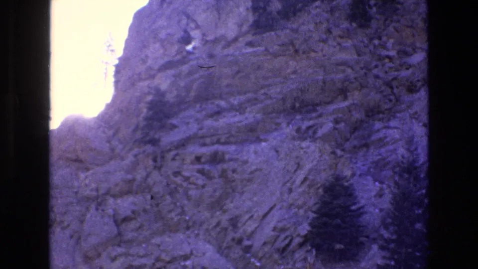 1973 Colorado 8mm frame showing rugged mountain rock, dark evergreen trees rooted in the slope, and strong highlight bloom near the upper-left edge.