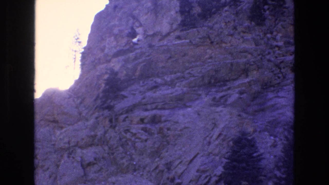 A white mountain goat appears small against a steep layered rock face in Colorado 1973 home-movie footage, with bright sky along the upper left edge and dark film borders visible.