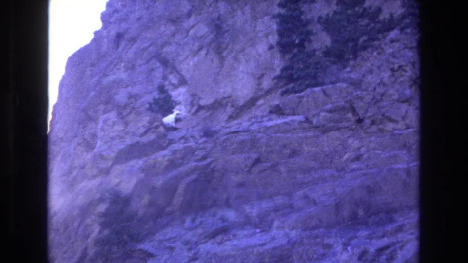 1973 Colorado 8mm frame showing a rugged purple-blue rocky cliff face, bright overexposed light at the left edge, and dark film borders around the image.