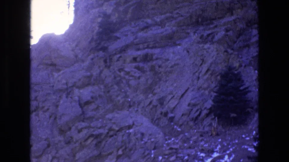 1973 Colorado 8mm frame showing a descending rocky slope, sparse evergreen trees, and a blown-out bright area along the left side of the frame.