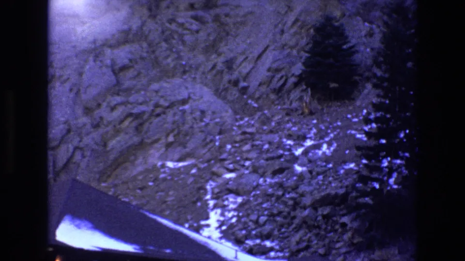 1973 Colorado 8mm frame showing a rocky mountain slope with evergreen trees, pale snow-like or reflective patches, and a dark angular structure near the lower edge.