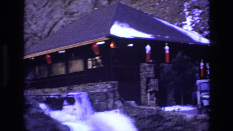 1973 Colorado 8mm frame showing a closer visitor view of the rustic lodge, including snow in front, stone pillars, red lanterns, dark siding, and the edge of a parked Jeep-like vehicle.