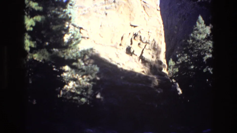 1973 Colorado home-movie frame showing a sunlit rocky cliff face bordered by evergreen trees, with a dark shadowed foreground suggesting the surrounding mountain terrain.