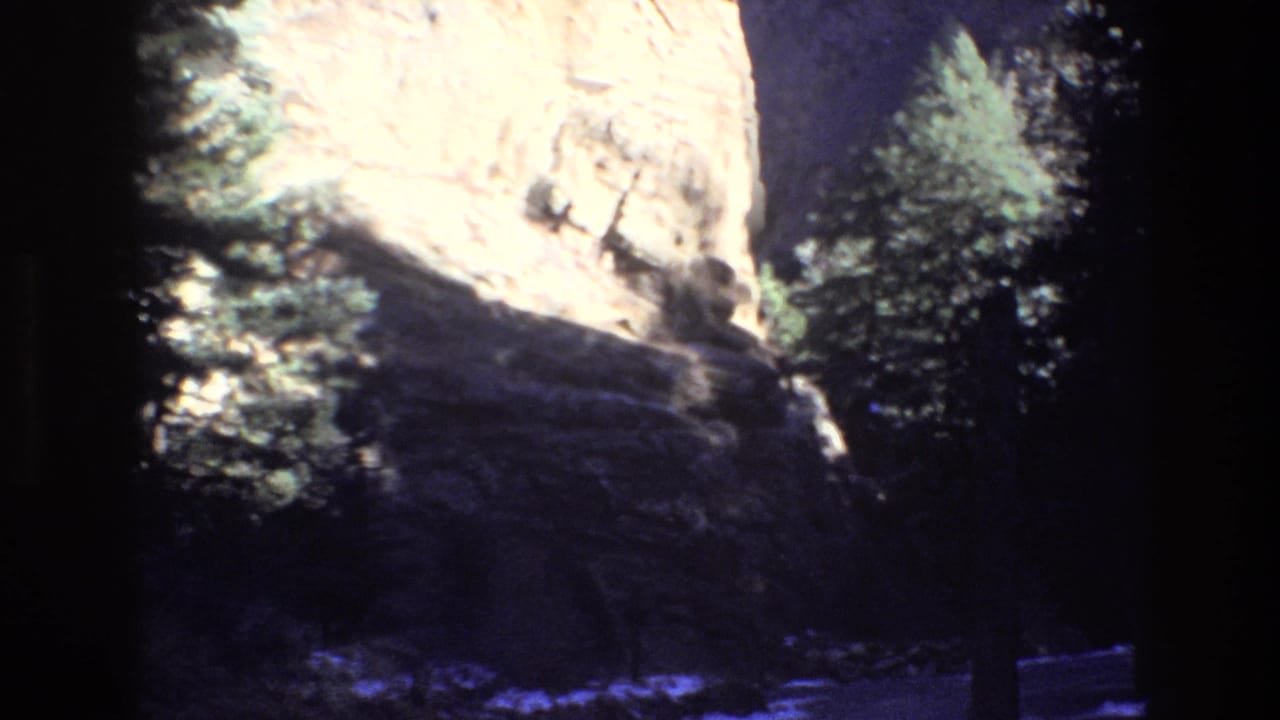 1973 Colorado 8mm frame showing a bright rock formation above dark foreground rocks and evergreen trees in deep shade.
