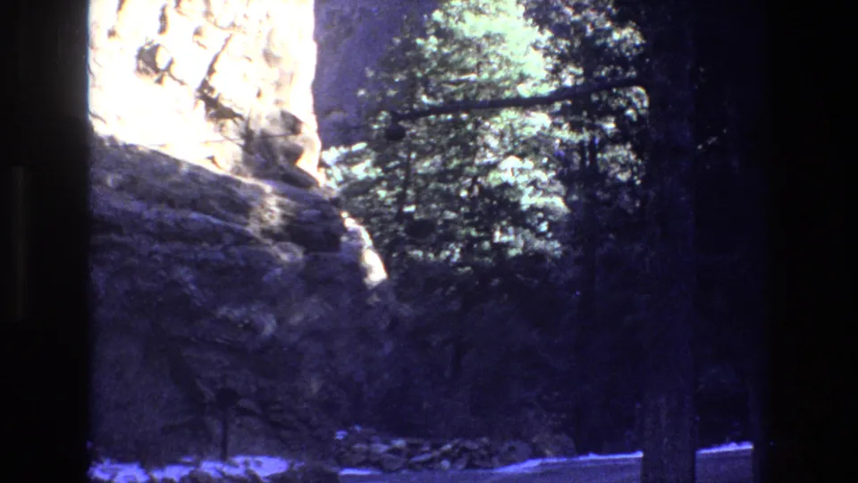 1973 Colorado 8mm frame showing a shaded boulder mass below a sunlit rock wall, with dense trees along the right side.