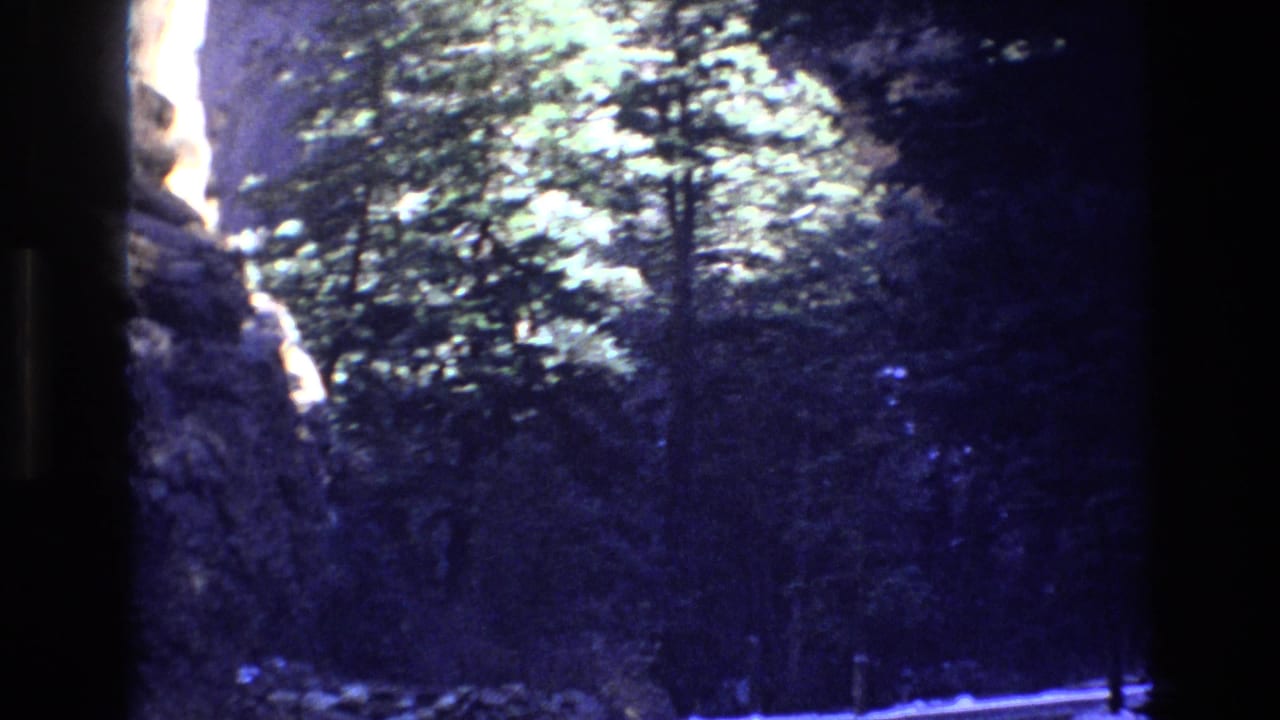 1973 Colorado home-movie frame showing a narrow road through evergreen forest with snow on the ground and rocky terrain at the edge.