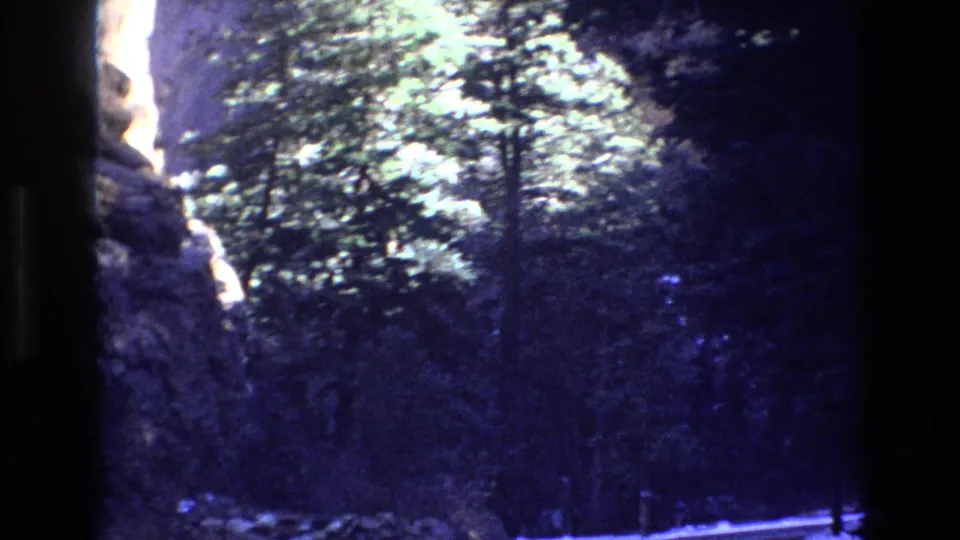1973 Colorado home-movie frame showing a narrow road through evergreen forest with snow on the ground and rocky terrain at the edge.