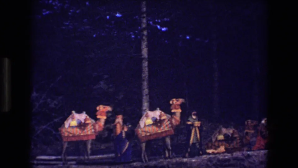 1973 Colorado home-movie frame showing a nighttime outdoor nativity-style scene with camels, robed figures, a tripod-like structure, and trees in the background.