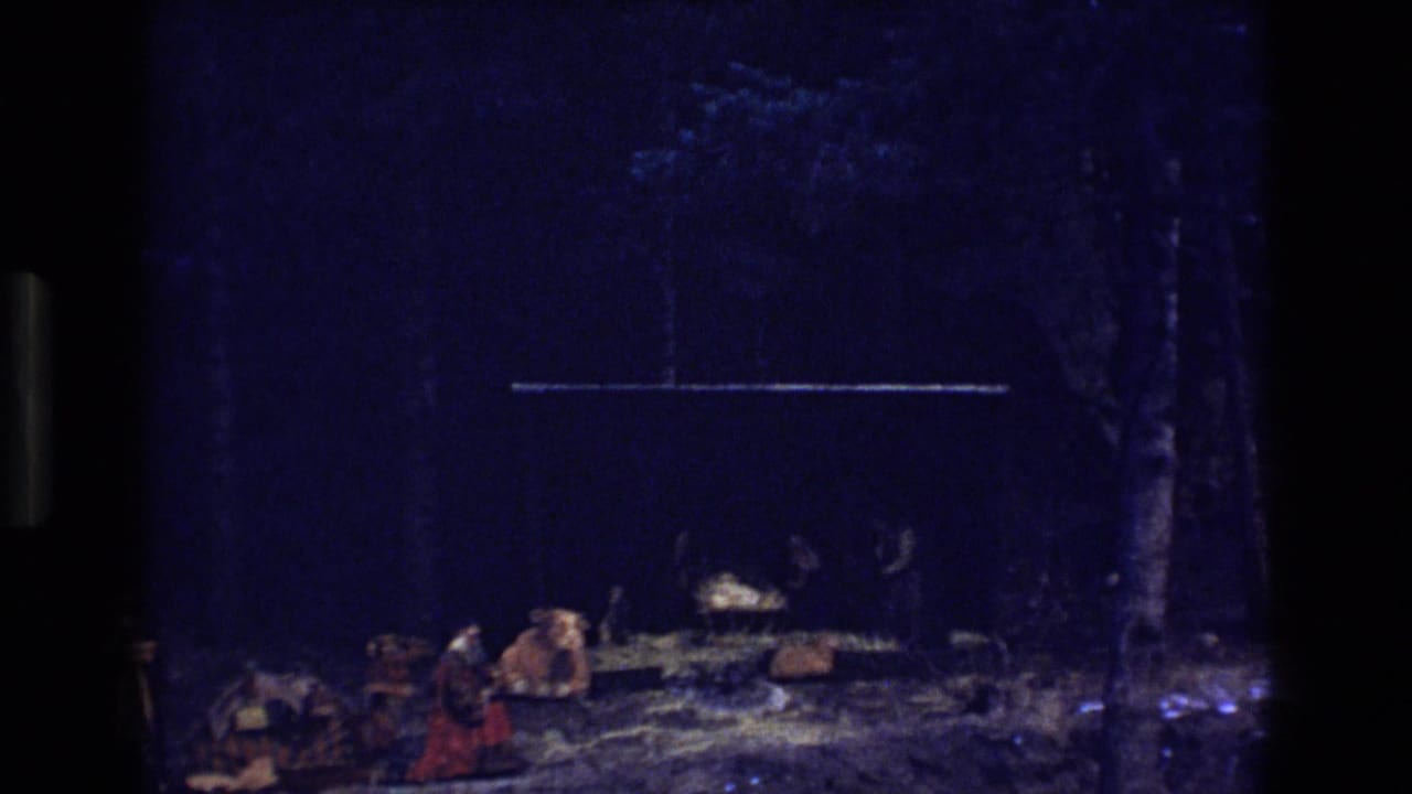 1973 Colorado 8mm frame showing a dimly lit nativity setup with a manger, straw, a cow figure, and people or costumed figures near the scene.