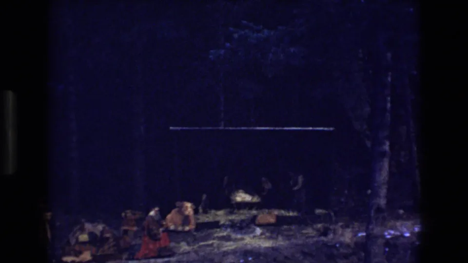 1973 Colorado 8mm frame showing a dimly lit nativity setup with a manger, straw, a cow figure, and people or costumed figures near the scene.