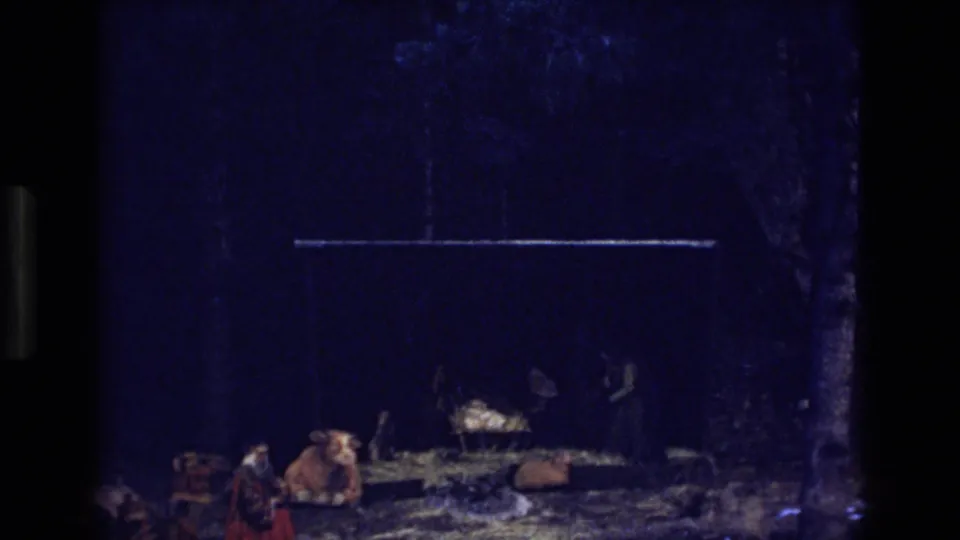 1973 Colorado 8mm frame of a dark nativity display, with a cow figure in front, straw around a manger, and indistinct figures standing near the cradle.