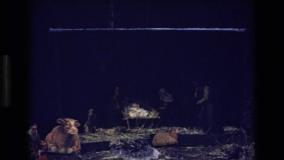 1973 Colorado 8mm frame of the nativity setting with a manger or cradle, straw, a cow figure, a reclining animal form, and indistinct people or figures in dark clothing.
