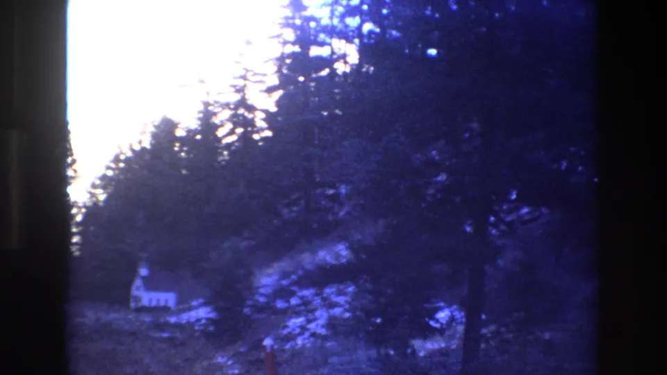 Overexposed 1973 Colorado 8mm frame showing a white house partly obscured by evergreen trees on a snowy hillside.