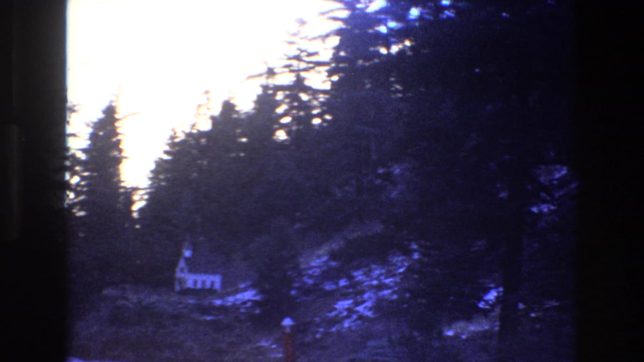 1973 Colorado 8mm frame showing a small white chapel nestled in shadowed evergreens with a pale cliff rising behind it.