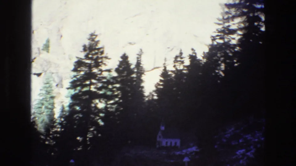1973 Colorado 8mm frame with pine silhouettes, a bright rocky cliff, and a small white chapel set low in the landscape.