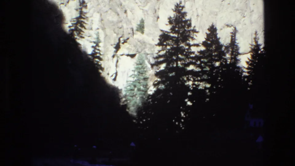 1973 Colorado 8mm frame emphasizing tall evergreens and a bright cliff face, with the chapel mostly obscured by trees and shadow.