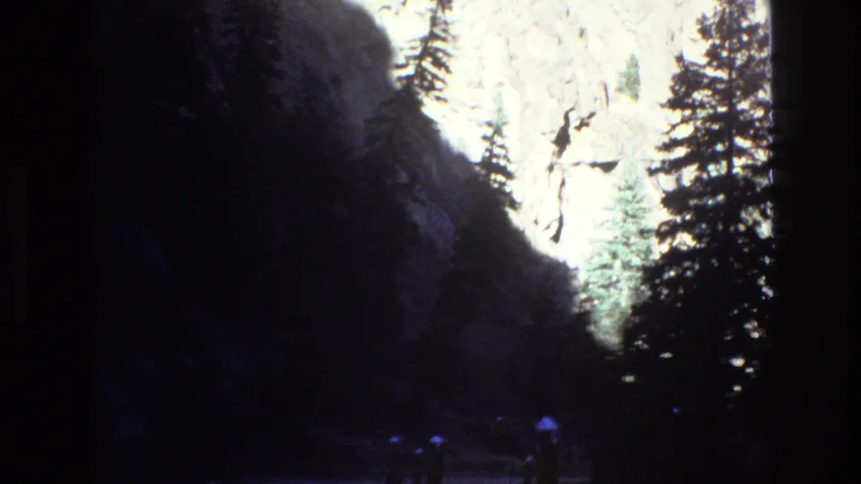 1973 Colorado 8mm frame showing a shadowed mountain scene with evergreen trees, a bright cliff, and small distant visitors near the lower edge.