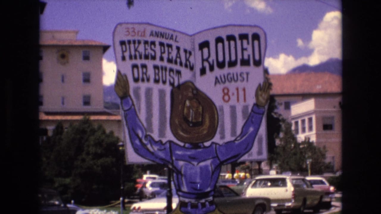 Large Pikes Peak Rodeo sign with cartoon cowboy, parked cars, buildings, sky, and mountains.