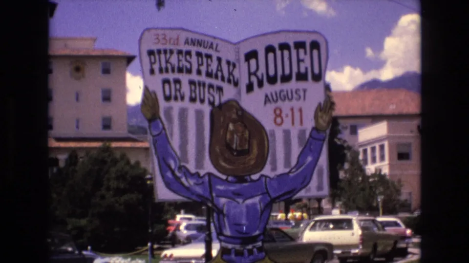 1973 Colorado Springs view of a large 33rd Annual Pikes Peak Rodeo sign with a cartoon cowboy, parked cars, buildings, blue sky, clouds, and distant mountains.