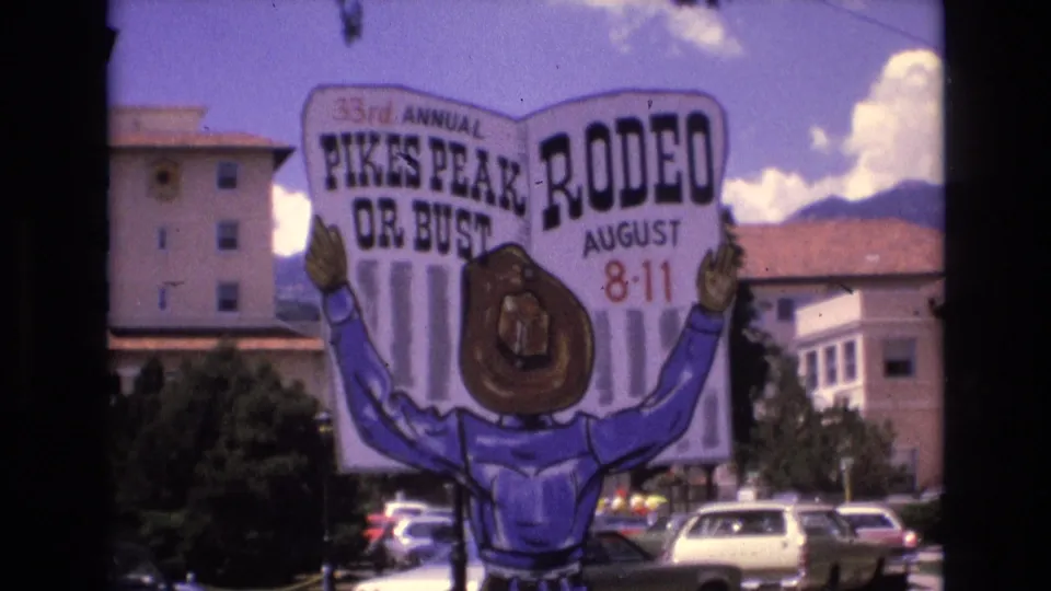 1973 Colorado Springs frame showing the Pikes Peak Rodeo or Bust sign with August dates, a cartoon cowboy, parked cars, buildings, clouds, and mountain backdrop.