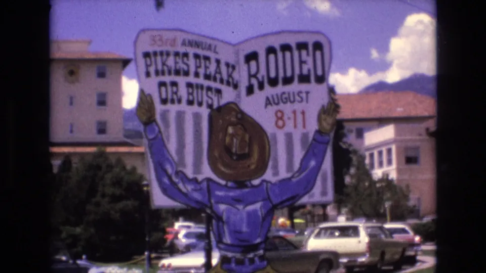 1973 Colorado Springs frame of the 33rd Annual Pikes Peak Rodeo sign with cartoon cowboy artwork, parked cars, nearby buildings, trees, clouds, and mountains behind it.