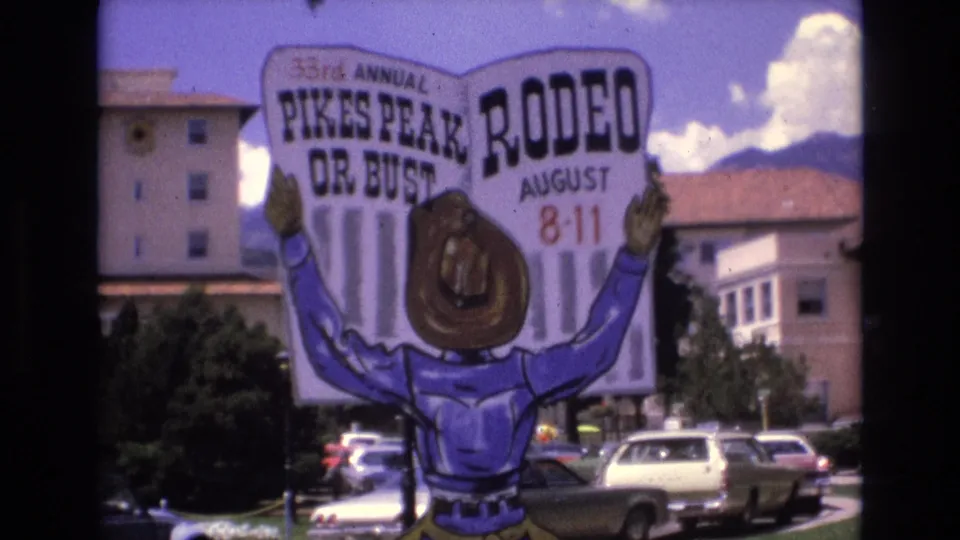 1973 Colorado Springs 8mm frame showing a colorful cowboy-shaped sign for the 33rd Annual Pikes Peak Rodeo, August 8-11, with parked cars, trees, and buildings in the background.