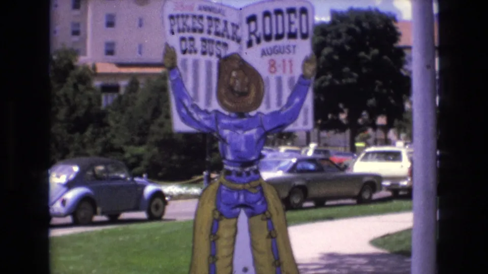 1973 Colorado Springs 8mm frame showing the Pikes Peak Rodeo sign from a slightly lower composition, with its cowboy legs visible above a lawn and parked 1970s cars.