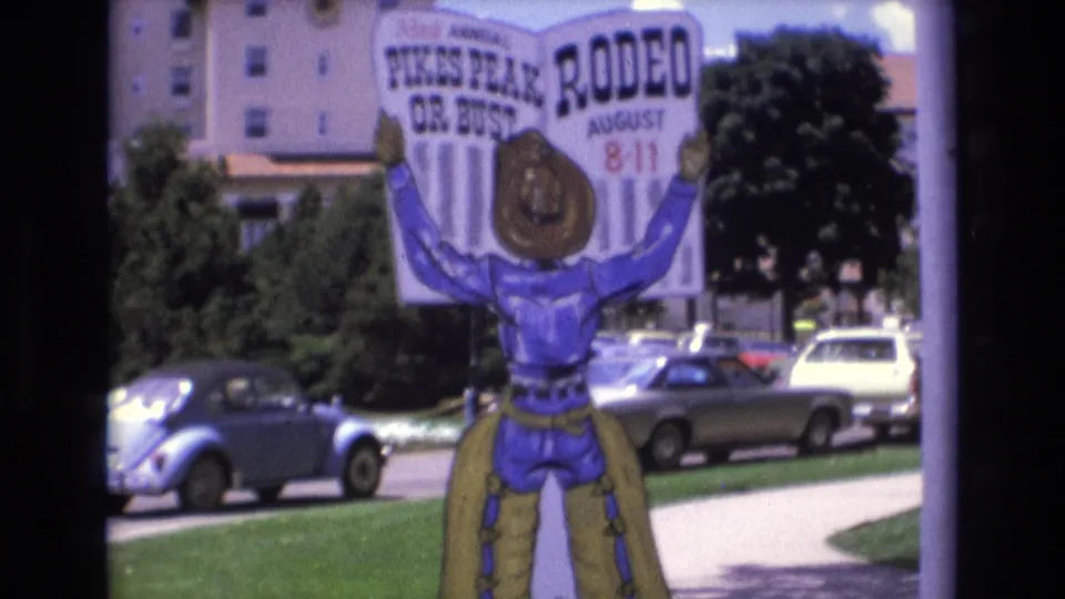 1973 Colorado Springs home-movie frame showing the Pikes Peak Rodeo sign against a lawn and curbside parking, with 1970s vehicles and trees framing the background.