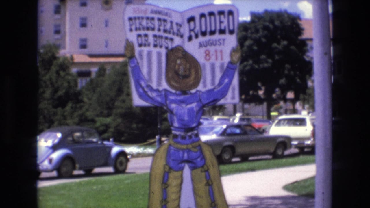 1973 Colorado Springs home-movie frame showing a large Pikes Peak or Bust Rodeo sign with a cowboy figure, parked cars, grass, trees, and a building behind it.